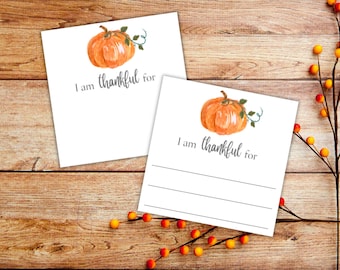 May include: Two white cards with a pumpkin illustration and the text "I am thankful for". The cards are on a wooden surface with a branch of berries.