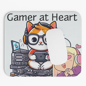 May include: A mouse pad with a white background and the text "Gamer at Heart" in black. A cartoon cat wearing headphones and glasses is holding a video game controller. A cartoon girl with pink hair is wearing headphones and looking at the cat.