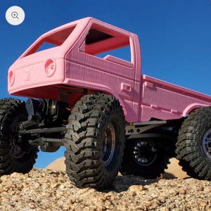 Könnte beinhalten: Ein rosa ferngesteuerter Truck mit großen, schwarzen Offroad-Reifen. Der Truck hat eine detaillierte Kabine und Ladefläche, vor blauem Himmel und felsigem Gelände. Das Fahrzeug ist für den robusten Außeneinsatz konzipiert.
