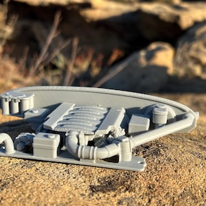 May include: A light gray model of an engine, featuring detailed components such as the intake manifold, exhaust pipes, and various other engine parts. The model is set against a blurred, natural background.