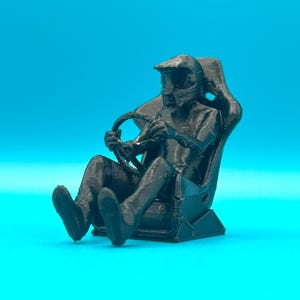 May include: A black 3D-printed figurine of a race car driver seated in a racing seat, holding a steering wheel. The driver wears a helmet. The figurine is set against a blue background.