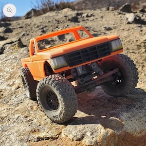 以下が含まれることがあります： オレンジ色のラジコンおもちゃのトラック。 大きく頑丈なタイヤが付いています。 トラックは、詳細なフロントグリル、ヘッドライト、キャブデザインを備えています。 岩の多い屋外の地形に配置されており、オフロードでの遊びを連想させます。