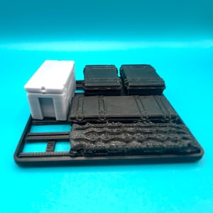 Könnte beinhalten: Eine Sammlung von kleinen, 3D-gedruckten Aufbewahrungsbehältern in verschiedenen Formen. Enthält eine weiße rechteckige Box mit einer vertieften Öffnung, mehrere schwarze Koffer und eine strukturierte schwarze Basis. Die Artikel sind auf einer schwarzen Plattform angeordnet.