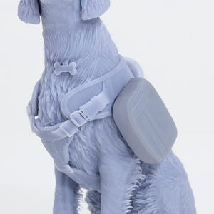 Puede incluir: Figura azul claro de un perro sentado, con un arnés con un detalle en forma de hueso y una pequeña mochila. El perro tiene pelo detallado y está sobre un fondo blanco. Probablemente un artículo decorativo.