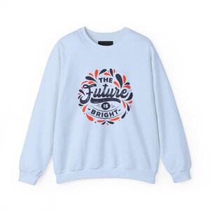 May include: Light blue sweatshirt with long sleeves. The front features a circular design with the words "The Future Is Bright" in navy blue and orange. The design is surrounded by orange and navy blue decorative elements.