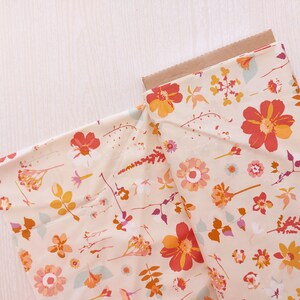May include: A roll of floral fabric with a cream backdrop, adorned with a pattern of orange, red, and yellow flowers, and green and purple leaves. The fabric is partially unrolled, showing a brown cardboard core.
