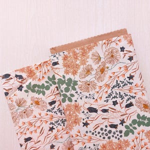 May include: Folded fabric with a floral design in orange, white, green, and navy blue. The fabric is partially inside a brown paper bag with a scalloped edge. The background is a light wood surface.