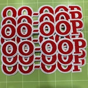 Peut inclure: Une collection d'écussons brodés présentant le texte "OO-OOP" en lettres rouges audacieuses avec une bordure blanche. Chaque écusson est construit avec une texture de fil rouge épais et surélevé et un contour blanc cousu, disposés en grille.