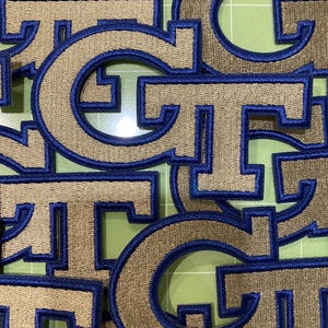 May include: A collection of embroidered patches featuring the letters G and T in a collegiate font. Each patch has a metallic gold centre with a thick, raised royal blue border. The letters are layered over one another in a dense, overlapping arrangement.