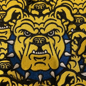 May include: A collection of embroidered patches featuring a yellow bulldog head with a scowling expression. The bulldog wears a blue spiked collar with silver-colored spikes. The design is rendered in yellow, black, blue, and silver thread.