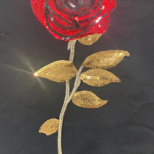 May include: A decorative rose applique featuring a vibrant red flower head constructed from dense, shimmering sequins. The stem and five distinct leaves are crafted from metallic gold sequins, providing a contrasting texture and color profile.