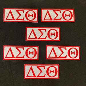 Peut inclure: Cinq écussons rectangulaires brodés présentant les lettres grecques Delta, Sigma et Theta en fil rouge sur fond blanc. Chaque écusson est bordé d'un contour rouge, et les lettres sont disposées horizontalement en motif répétitif.