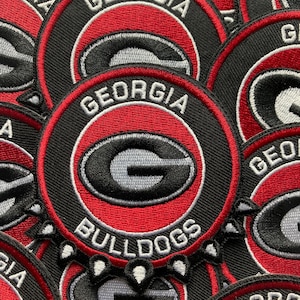 Puede incluir: Parches bordados circulares con el logotipo de los Georgia Bulldogs. Los parches tienen un borde negro, un círculo interior rojo y las palabras "GEORGIA" y "BULLDOGS" en blanco. El diseño central es una "G" gris sobre fondo negro.