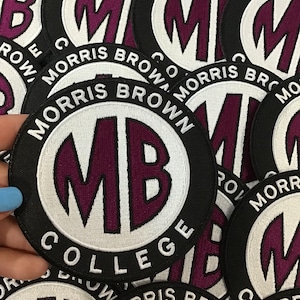 Pode incluir: Patches bordados circulares com o texto "MORRIS BROWN COLLEGE" em branco sobre fundo preto. O centro apresenta um oval branco com as letras "MB" em bordô.