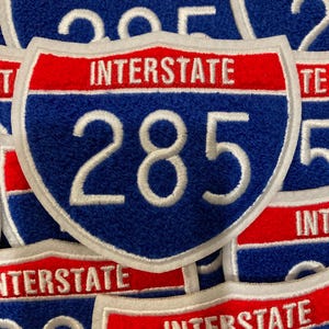 May include: A collection of shield-shaped embroidered patches featuring the text INTERSTATE in white on a red background above the number 285 in white on a blue background, all outlined with a white border, arranged in a layered overlapping stack.