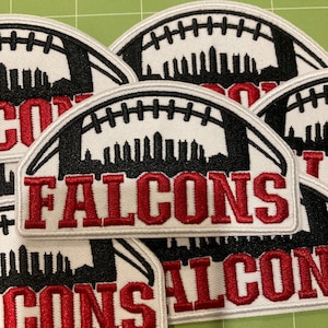 Pode incluir: Uma coleção de emblemas bordados com o formato de uma bola de futebol americano, uma silhueta preta de horizonte urbano e a palavra FALCONS bordada em letras vermelhas. Os emblemas têm uma borda branca e estão dispostos em camadas.