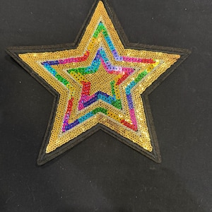 Könnte beinhalten: Ein sternförmiger Aufnäher mit einem zentralen goldenen Paillettenstern, der von einem mehrfarbigen Paillettenrand in Blau-, Grün-, Rosa- und Gelbtönen umgeben ist. Der gesamte Stern ist mit einem dicken schwarzen Stickrand auf schwarzem Stoff.