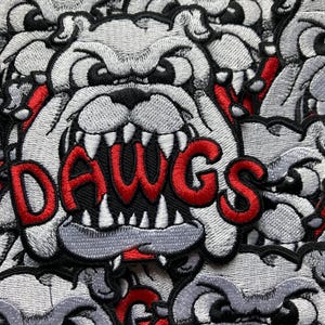 May include: Close-up of embroidered patches featuring a bulldog design. The patches are primarily gray, black, and red. The word "DAWGS" is embroidered in red across the bulldog's mouth. The patches are densely packed together.