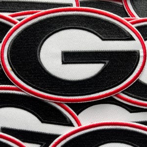 Könnte beinhalten: Nahaufnahme von mehreren gestickten Aufnähern. Jeder Aufnäher zeigt einen schwarzen Buchstaben "G" auf weißem Grund, umrandet von Rot. Die Aufnäher sind gestapelt und zeigen das Design und die Stoffstruktur.