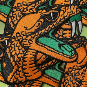 May include: A collection of embroidered patches featuring a snake head design. The snake is orange with black scale detailing, a bright green inner mouth, and sharp white fangs. The design is outlined in black thread with a bold, graphic style.