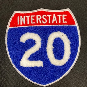 May include: A shield-shaped patch featuring the text INTERSTATE in white on a red background at the top, with the number 20 in white on a blue textured background below. The entire patch is outlined with a thin white border.