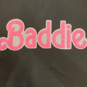 May include: A black fabric surface featuring the word Baddie in a bright pink, textured, fuzzy, chenille-style font. The letters have a light gray outline, creating a distinct contrast against the dark background of the material.