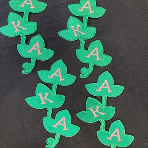 以下が含まれることがあります： 3枚の葉を持つ緑色のアイビーのつるの形をした刺繍ワッペン4枚セット。各ワッペンには、ピンク色のギリシャ文字「A」「K」「A」が葉の上に縦に刺繍されています。ワッペンは質感のある仕上がりで、緑色の縁取りが施されています。