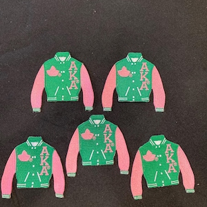 Può includere: Cinque toppe ricamate a forma di giacche varsity. Ogni giacca è verde con maniche rosa, presenta un emblema a foglia di edera rosa sul petto e le lettere AKA impilate verticalmente in rosa. Le toppe hanno dettagli a bottone bianchi.
