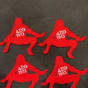 Puede incluir: Cuatro parches bordados rojos idénticos, cada uno con forma de figura en cuclillas. El centro de cada parche presenta las letras griegas blancas Delta Sigma Theta y el número 1913, cosidos directamente sobre la superficie de tela roja.