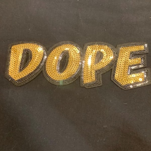 May include: A decorative patch featuring the word DOPE in bold, capital letters. The letters are covered in shimmering gold sequins and are outlined with a thick, black fabric border, creating a high-contrast, textured appearance.