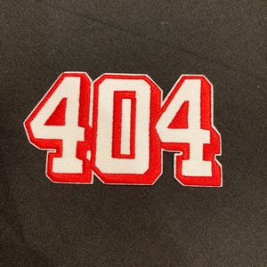 May include: A black fabric surface featuring an embroidered patch with the number 404. The digits are rendered in a bold, white block font with a thick, vibrant red outline, creating a distinct three-dimensional appearance against the dark background.