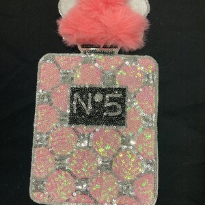 Puede incluir: Un parche rectangular con forma de frasco de perfume, cubierto de lentejuelas rosas y plateadas en un patrón de rejilla. Un centro rectangular negro presenta el texto N°5 en plateado. Un pompón rosa esponjoso corona el cuello del frasco.