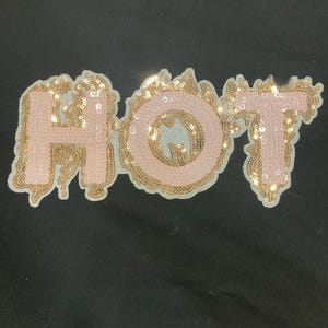 May include: A fabric patch featuring the word HOT in capital letters. The letters are filled with light pink sequins, while the surrounding flame-like border is crafted from shimmering gold sequins, creating a distinct contrast against the background.