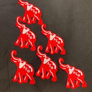 Peut inclure: Cinq écussons brodés en forme d'éléphant rouge, chacun présentant un contour blanc et les lettres grecques Delta Sigma Theta en blanc. Les motifs d'éléphants sont orientés avec les trompes levées, affichant une texture brodée cohérente.