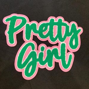 May include: A fabric patch featuring the text Pretty Girl in a cursive font. The letters are green with a thick pink border and a white outline, creating a layered effect. The patch is set against a solid black background, showcasing the vibrant colors.
