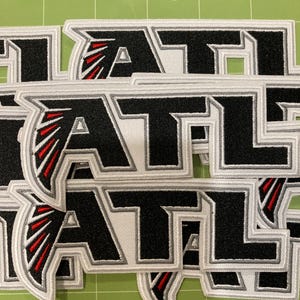 Könnte beinhalten: Eine Sammlung von gestickten Aufnähern mit den Buchstaben ATL in fetter schwarzer Schrift mit weißem und grauem Rand. Der Buchstabe A enthält eine stilisierte rot-schwarze Flügelgrafik. Die Aufnäher sind in einem Stapel übereinandergelegt.