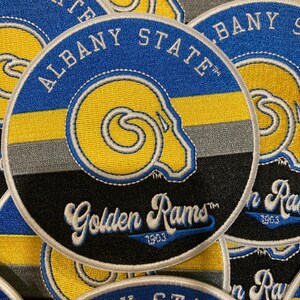 May include: A circular embroidered patch featuring the text Albany State at the top and Golden Rams 1903 at the bottom. The design includes a yellow ram head logo centred over horizontal blue, yellow, and grey stripes on a black background.