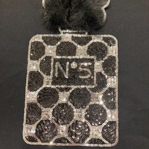 Puede incluir: Un parche rectangular con forma de frasco de perfume, cubierto de lentejuelas negras y plateadas dispuestas en un patrón de panal. El centro presenta el texto N°5 en lentejuelas plateadas y un pompón de piel sintética negra en la parte superior.