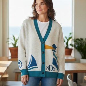 May include: Cream-colored cardigan sweater with teal trim and button closure. The sweater features nautical-themed designs, including a sailboat, lighthouse, and the initials "sDs". The sweater is worn over a white t-shirt.