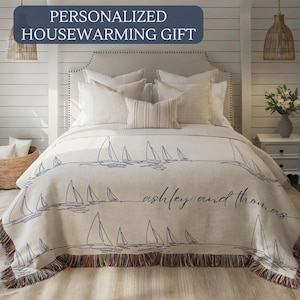 May include: A cream-colored throw blanket with a sailboat design and the names "ashley and thomas" in script. The blanket has a fringed edge with blue, red, and brown threads. The text "PERSONALIZED HOUSEWARMING GIFT" is at the top.