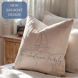 May include: Beige linen pillow with a sailboat design and the text "The Anderson Family." The pillow features a new sailboat design and is placed on a bed with other pillows. The pillow is a decorative item.