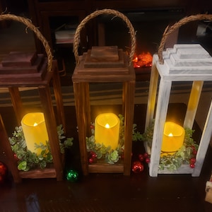 May include: Three wooden lanterns in varying shades of brown and white, each with a rope handle. Inside each lantern is a glowing candle surrounded by greenery and red berries. Decorative Christmas ornaments are scattered around the base.