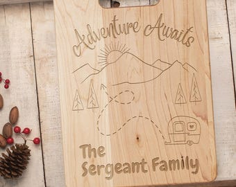 May include: A wooden cutting board with an engraved design of a camper driving through a mountain landscape. The text "Adventure Awaits" and "The Sergeant Family" is engraved on the board.