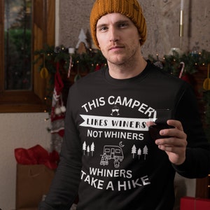 May include: Black sweatshirt with white text that reads "This Camper Likes Winers Not Whiners Whiners Take A Hike". The text is surrounded by a camper icon, wine glasses, and pine trees. A person is wearing the sweatshirt and holding a glass of red wine.