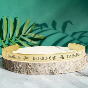 May include: Gold-toned metal bracelet with the inscription "Breathe In ∞ Breathe Out ∞ I've got this" engraved on it.