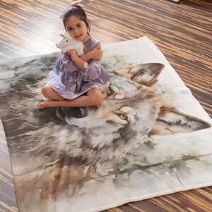 May include: A white fleece blanket with a watercolor painting of a wolf's face. A young girl is sitting on the blanket, holding a white stuffed unicorn.