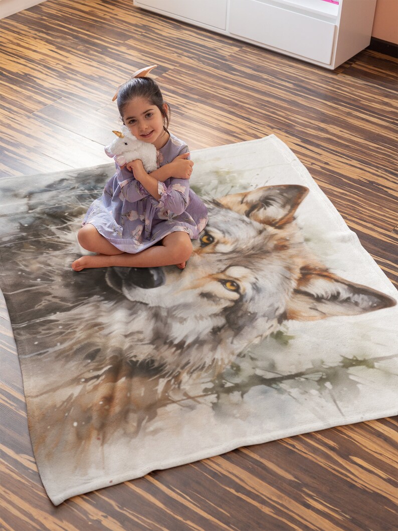 May include: A white fleece blanket with a watercolor painting of a wolf's face. A young girl is sitting on the blanket, holding a white stuffed unicorn.