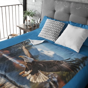 May include: A blue fleece blanket featuring a printed image of a bald eagle in flight against a cloudy sky. The eagle is brown and white with its wings outstretched. The blanket is on a blue bed with white and grey pillows.