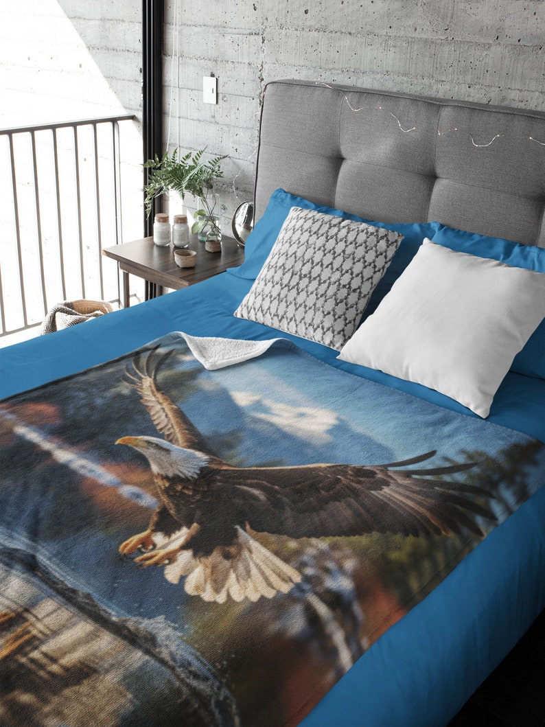 May include: A blue fleece blanket featuring a printed image of a bald eagle in flight against a cloudy sky. The eagle is brown and white with its wings outstretched. The blanket is on a blue bed with white and grey pillows.