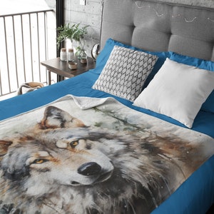 May include: A soft, blue fleece throw blanket with a watercolor painting of a wolf's face. The wolf has brown fur, yellow eyes, and a black nose. The background is a blurry watercolor of trees and leaves.
